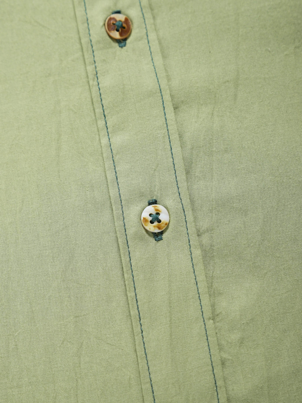 Close-up of pale olive plant-dyed organic cotton shirt placket with natural shell buttons and contrast stitching.