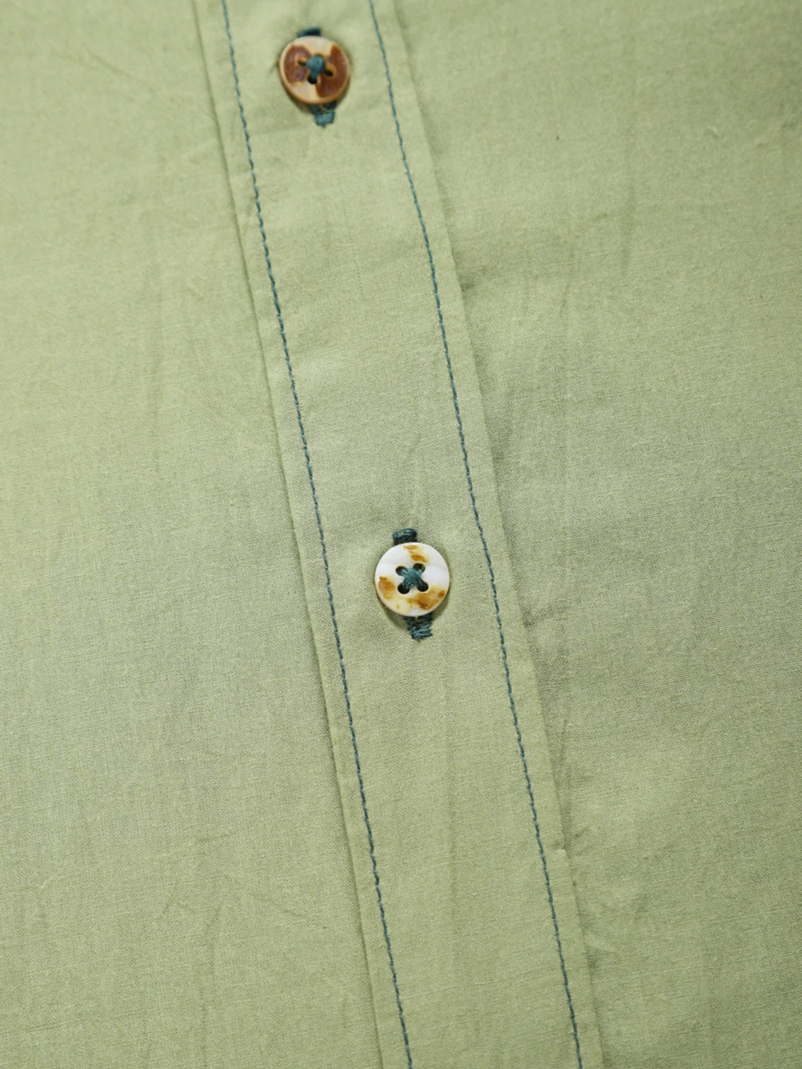 Close-up of pale olive plant-dyed organic cotton shirt placket with natural shell buttons and contrast stitching.